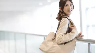 Woman smiling and standing by a railing with a beige bag.